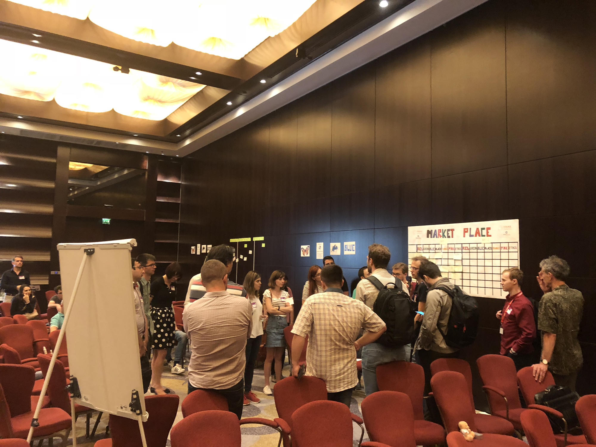 Attendees standing in small groups around a hand-drawn marketplace board covered in sticky notes, choosing their own sessions