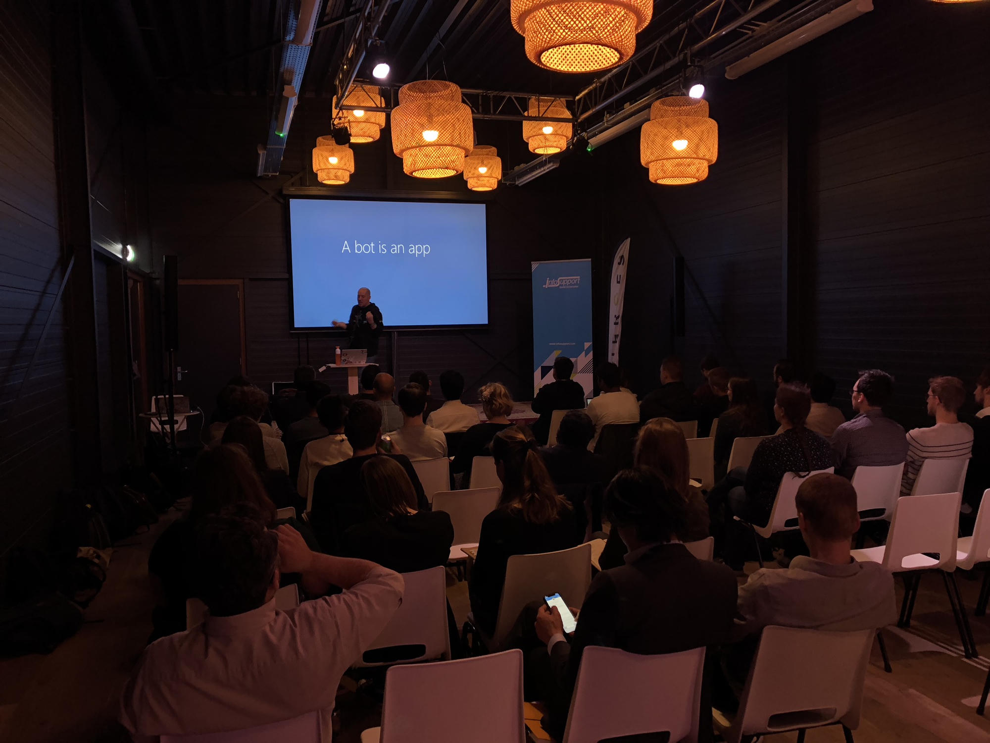A meetup audience seen from the back of a dark room, rows of people watching a speaker, one of them reading something on their phone