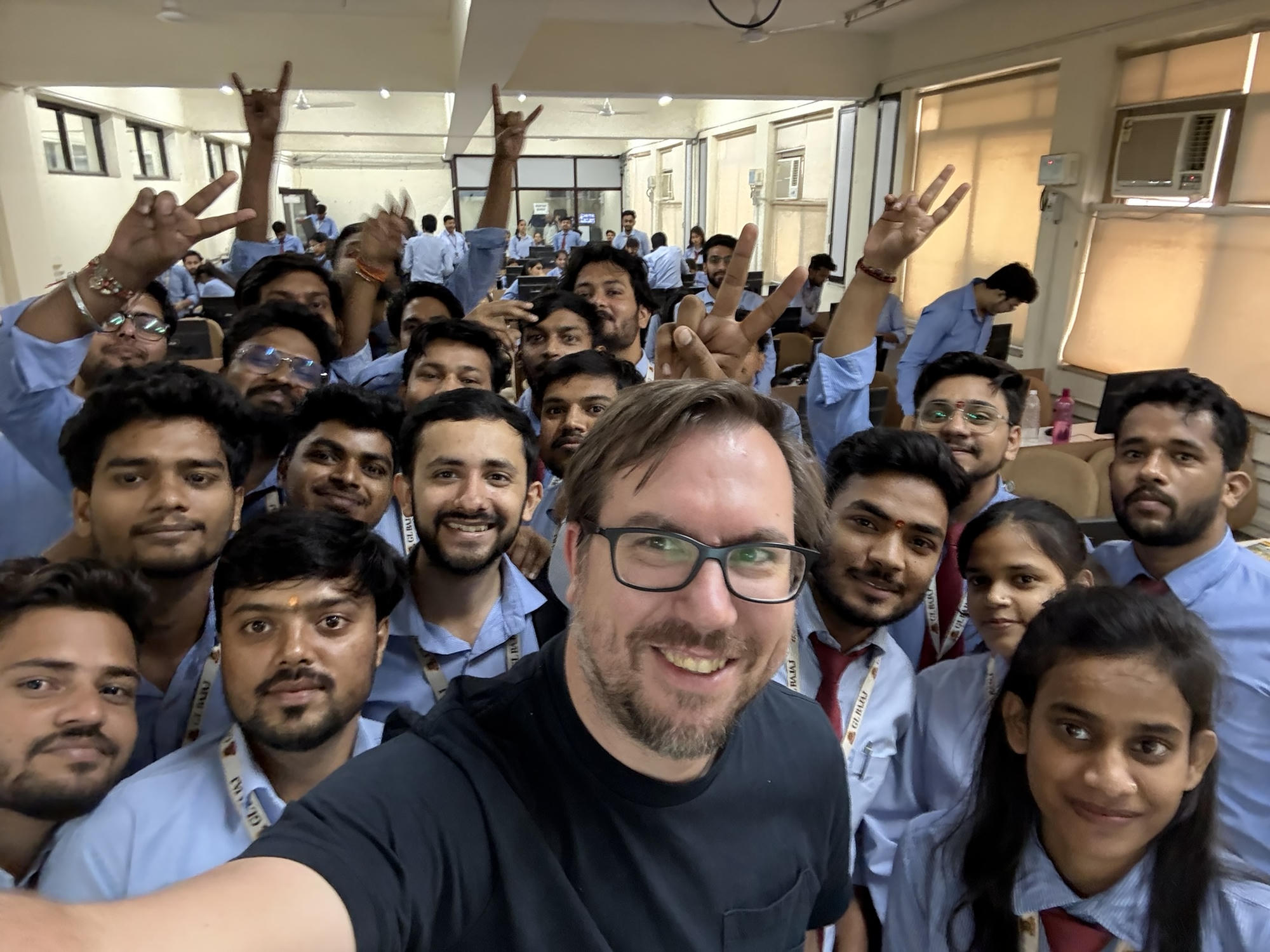 A packed university computer lab in India, students in blue uniform shirts crowding into a selfie with me, hands up in peace signs and rock signs, more of them still at their machines in the rows behind