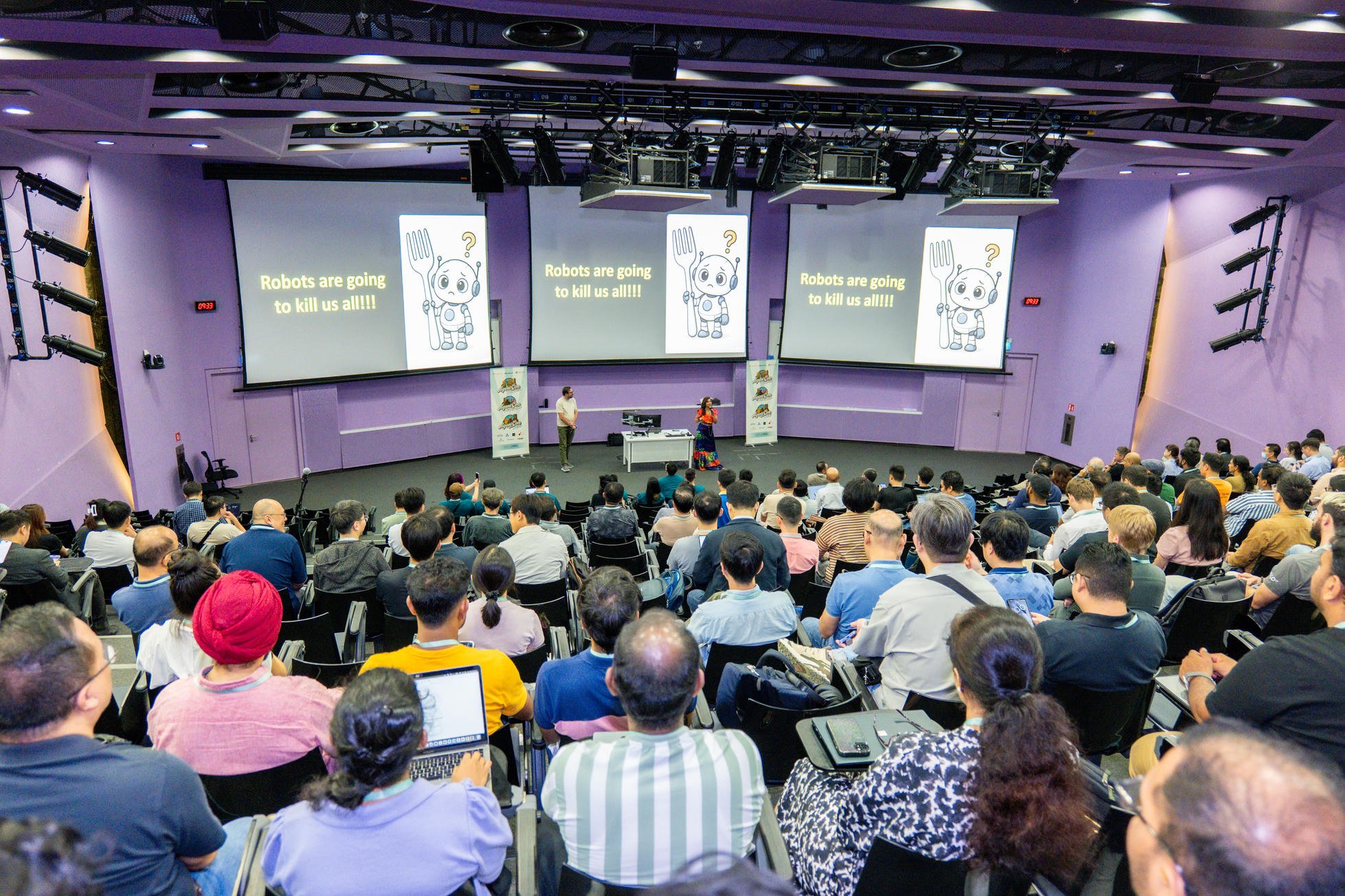 A packed auditorium seen from the back, with two presenters on stage and three large screens showing their talk