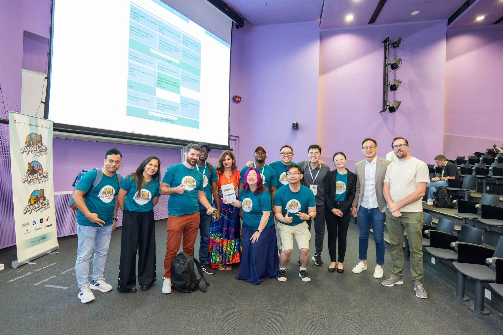 A group posing together in an auditorium, several wearing teal AgentCon shirts, with Henk on the right