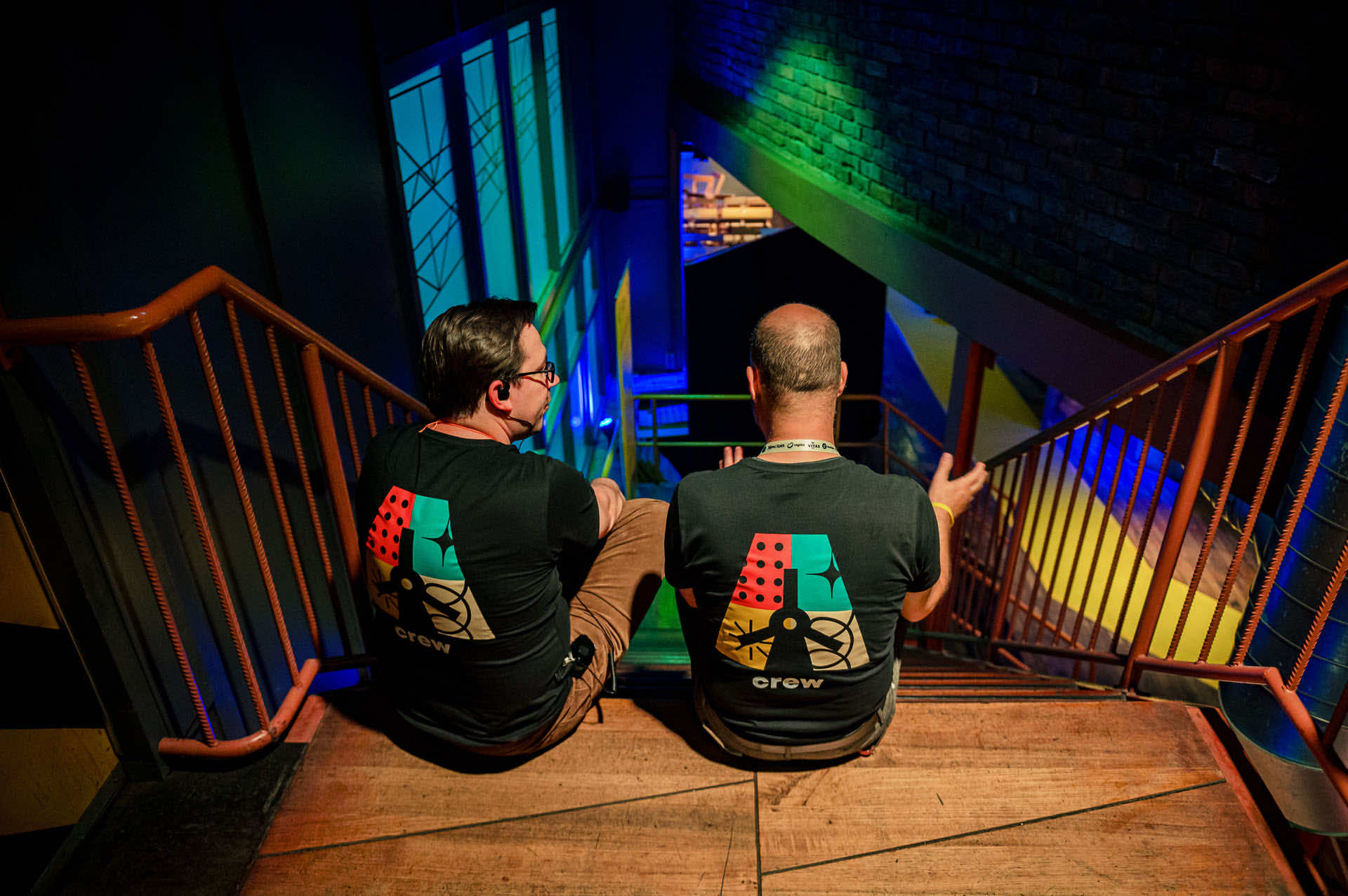 Two crew members in black crew shirts sitting on a stairwell above the venue, backs to the camera, looking out over the blue and green stage lighting below
